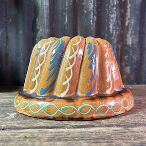 May include: A brown ceramic bundt cake pan with a decorative design of blue, white, and green floral patterns. The pan has a fluted top and a smooth bottom.