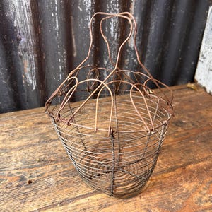 May include: A vintage wire basket with a handle. The basket is made of rusty metal and has a rounded shape. The handle is formed by two curved wires that meet at the top. The basket is likely used for gathering eggs or produce.