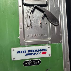 May include: Close-up of a green metal panel with an Air France logo and the text "AIR FRANCE" in capital letters. A silver metal latch is visible, with "OPEN" and "CLOSE" indicators. The panel also has the number "VP0052".