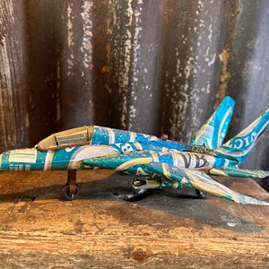 Puede incluir: Un avión modelo hecho de latas de aluminio recicladas. El avión está pintado en tonos azules y verdes, con una cabina marrón. El avión está sentado sobre una superficie de madera.