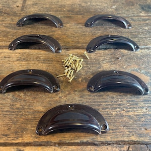 May include: Seven dark brown, semi-circular cup drawer pulls with a smooth, glossy finish. Each pull features two mounting holes at the ends. A small pile of gold-coloured metal screws is positioned in the centre of the arrangement of pulls.