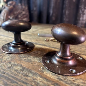 May include: Two oval-shaped door knobs with a dark bronze finish. Each knob features a smooth, rounded handle attached to a circular mounting plate with three visible screw holes. The metal surface displays a warm, metallic brown patina.