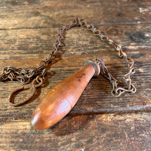 May include: A vintage wooden pull handle with a metal chain and hook. The handle is a warm brown colour, and the chain and hook show signs of age and rust. The item is likely a part of a light fitting or similar mechanism.