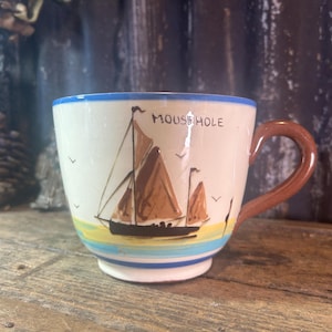 May include: A ceramic mug with a brown handle and a blue rim. The mug features a hand-painted sailboat scene with the text "MOUSEHOLE". The design includes brown sails, a black boat, and a yellow and blue water line.
