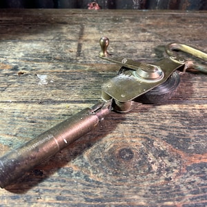 Puede incluir: Cinta métrica antigua de latón con manivela y asa. La cinta métrica está enrollada dentro de la carcasa de latón. La cinta métrica está sobre una superficie de madera desgastada.