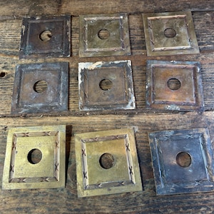 Puede incluir: Nueve placas metálicas cuadradas antiguas con un agujero central, dispuestas sobre una superficie de madera. Las placas varían en color, desde bronce hasta oro, con bordes decorativos. Algunas muestran signos de edad y desgaste.