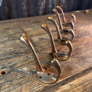 May include: A collection of five antique bronze-toned metal coat hooks. Each hook features a curved design with a rounded end and a base with screw holes. They are displayed on a weathered wooden surface.