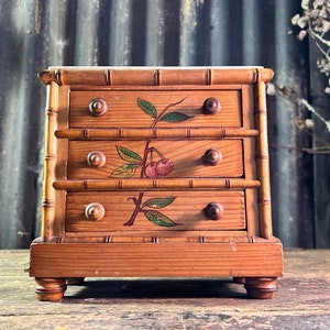 May include: A small, wooden chest of drawers with three drawers. The chest is painted with cherry branch designs. The wood has a bamboo-like design on the sides. The chest sits on four small, rounded feet.