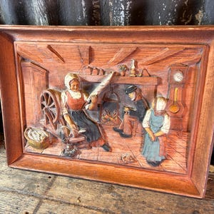 以下が含まれることがあります： 3人の人物がいる家庭的なシーンを描いた、額入りの彫刻された木製レリーフ。女性が糸車で糸を紡ぎ、他の2人は暖炉の近くにいます。レリーフは茶色の木製フレームに入っています。