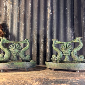 Puede incluir: Un par de raspadores de botas de hierro fundido antiguos con una pátina verde. Cada raspador presenta un par de caballitos de mar estilizados que flanquean una concha de vieira.