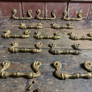 May include: A collection of 12 antique brass drawer pulls with a decorative, ornate design. The pulls are in the shape of a loop with a decorative scroll design.