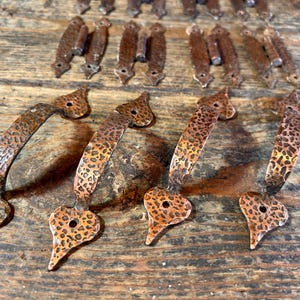 Op de afbeelding: Een verzameling antieke koperkleurige kasttrekkers en scharnieren. De trekkers hebben een getextureerd, gehamerd ontwerp met een hartvormige basis. De scharnieren zijn rechthoekig met decoratieve details.