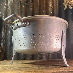 May include: A round, brushed aluminum colander featuring a wide rim, two sturdy side handles attached with rivets, and three curved metal legs. The body of the colander is perforated with small, evenly spaced holes for drainage.