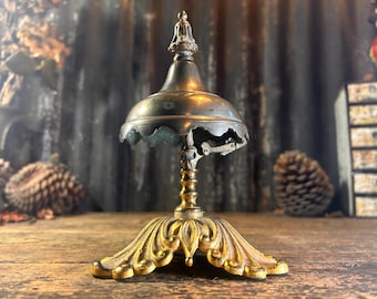 May include: A vintage brass hotel bell with a gold-toned base. The bell has a decorative, ornate design and a scalloped edge.