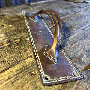 Könnte beinhalten: Ein rechteckiger Türgriff aus Messing mit einem dekorativen Rand und einem gebogenen Griff. Der Griff ist auf einer Holzoberfläche montiert.