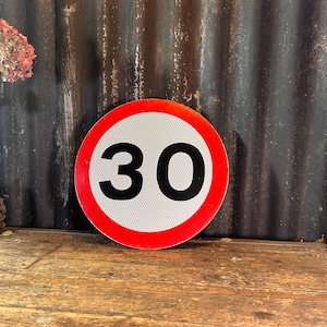 May include: A circular road sign featuring a white centre, a red border, and the number 30 in black. The sign is displayed on a wooden surface with a dark, corrugated metal background. Decorative elements include dried flowers and a pinecone.