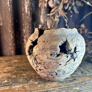 May include: A small, brown, ceramic pot with a rough, textured surface. The pot has several cracks and holes, giving it a rustic and aged appearance.