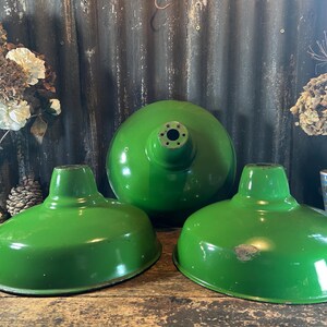 May include: Three green metal lampshades, one large and two smaller, on a wooden surface. The lampshades have a glossy finish. The background features a corrugated metal wall and decorative items.