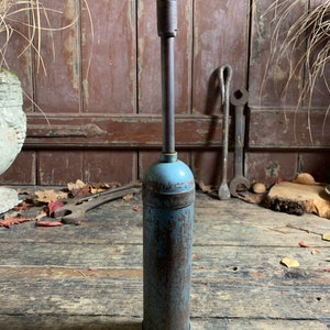 May include: A vintage blue and brown metal grease gun with a long nozzle. The grease gun is sitting on a wooden surface.