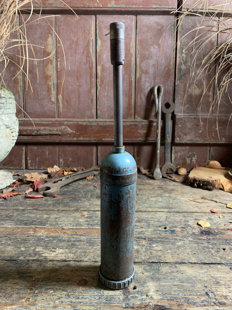 May include: A vintage blue and brown metal grease gun with a long nozzle. The grease gun is sitting on a wooden surface.