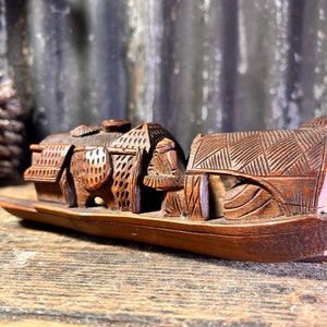 Op de afbeelding: Een met de hand gesneden houten sculptuur van een boot met een figuur die in de boeg staat. De boot is versierd met ingewikkelde houtsnijwerk en de figuur draagt een hoed. De sculptuur is gemaakt van donkerbruin hout en heeft een rustieke uitstraling.