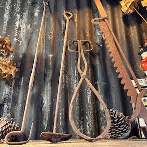 Op de afbeelding: Vier antieke roestige metalen haardgereedschappen die aan een gegolfde metalen muur hangen. De gereedschappen omvatten een poker, tang, een schep en een zaag. De gereedschappen zijn allemaal in verschillende staat van roest en slijtage.