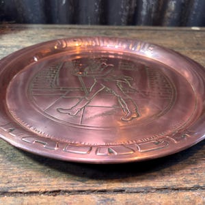 Puede incluir: Un plato de metal color cobre con un dise&ntilde;o en relieve. El plato presenta una imagen central de un hombre caminando con un bast&oacute;n, rodeado de texto. El plato tiene un borde elevado y se exhibe sobre una superficie de madera.