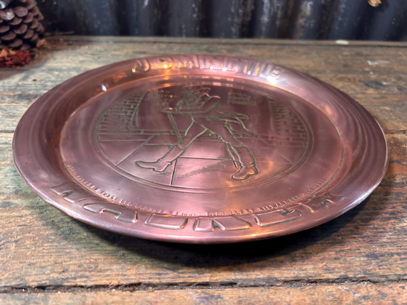 Puede incluir: Un plato de metal color cobre con un dise&ntilde;o en relieve. El plato presenta una imagen central de un hombre caminando con un bast&oacute;n, rodeado de texto. El plato tiene un borde elevado y se exhibe sobre una superficie de madera.