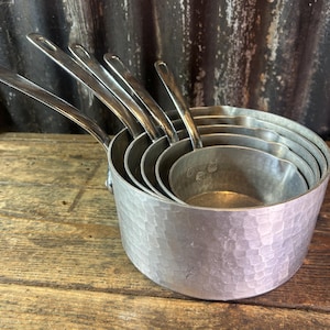 May include: A set of five nested silver-coloured metal saucepans with long handles. Each pan features a distinct hammered texture on the exterior surface and a smooth interior finish. The pans are stacked inside one another, showing graduated sizes.