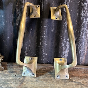 May include: Two matching brass door handles with a polished finish. Each handle features a long, curved grip and two rectangular mounting plates, each containing two pre-drilled screw holes for installation on a door or cabinet surface.