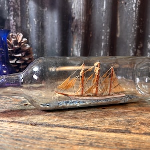 Puede incluir: Una botella de vidrio transparente que contiene un velero en miniatura. La botella tiene una tapa naranja y dorada. El velero tiene velas de color beige y está sobre una base azul y blanca, que representa el agua. La botella está sobre una superficie de madera.