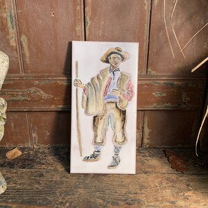 Può includere: Una piastrella di ceramica dipinta a mano che raffigura un uomo con un cappello e abiti tradizionali, che tiene un bastone da passeggio. La piastrella ha uno sfondo bianco ed è incorniciata da un bordo bianco.