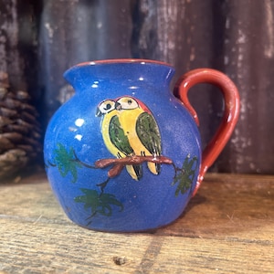 May include: A small, blue ceramic jug with a red handle and rim. The jug features a hand-painted design of two yellow and green parrots perched on a branch with green leaves. It sits on a rustic wooden surface.