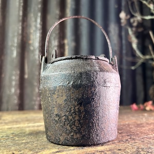 Op de afbeelding: Een kleine, rustieke gietijzeren pot met een handvat. De pot is donkerbruin met een versleten, verweerde uitstraling. Hij heeft een licht afgeronde vorm en een brede opening.