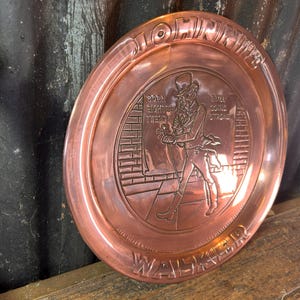 Puede incluir: Un plato decorativo redondo de color cobre con texto en relieve y la figura de un hombre con sombrero de copa. El plato tiene las palabras "JOHN WALKER", "BORN EIGHTEEN TWENTY" y "STILL GOING STRONG" en los bordes.