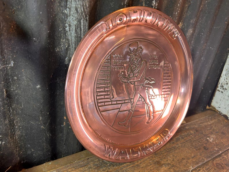 Puede incluir: Un plato decorativo redondo de color cobre con texto en relieve y la figura de un hombre con sombrero de copa. El plato tiene las palabras "JOHN WALKER", "BORN EIGHTEEN TWENTY" y "STILL GOING STRONG" en los bordes.