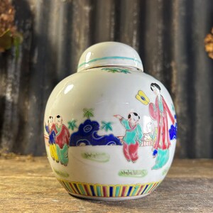 May include: White ceramic jar with a lid, decorated with colourful hand-painted figures of people in traditional clothing. The jar has a blue, yellow and red striped band around the bottom.
