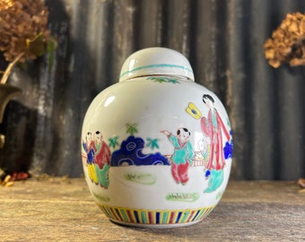 Puede incluir: Jarrón de cerámica blanca con tapa, decorado con figuras pintadas a mano de personas con ropa tradicional. El jarrón tiene una banda de rayas azules, amarillas y rojas alrededor de la base.