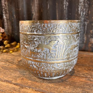 May include: A brass-coloured, cylindrical container with intricate etched designs. The container features a detailed relief of animals and foliage, with a band of gold-coloured metal at the top and bottom. The container sits on a wooden surface.
