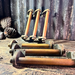 Puede incluir: Cuatro tiradores de puerta de latón antiguo con forma cilíndrica y extremo redondeado. Los tiradores están dispuestos en dos filas, la fila superior muestra los tiradores fijados a la pared y la fila inferior muestra los tiradores colocados planos sobre una superficie de madera.