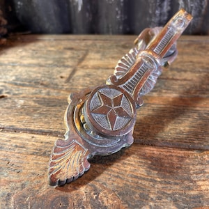 Könnte beinhalten: Ein antiker Schubladengriff aus Bronze mit einem Sternemblem. Die dekorative Hardware weist ein verziertes Design mit flügelartigen Details und einem zentralen Stern auf. Der Griff im antiken Stil ist auf einer rustikalen Holzoberfläche platziert.