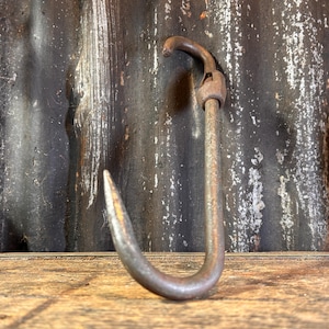 May include: A rusty metal hook with a sharp point and a loop at the top. The hook is hanging on a wooden surface with a corrugated metal background.