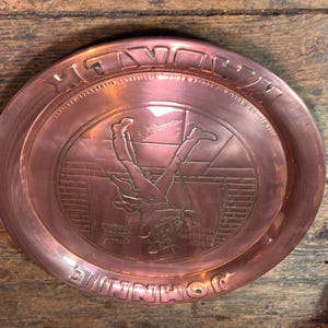 Puede incluir: Una bandeja de metal color cobre con un dise&ntilde;o en relieve. La bandeja presenta una imagen central de una figura cayendo, rodeada de texto. Las palabras "SHINNIHO" y "HE'S DOWN" son visibles. La bandeja es circular y tiene un borde elevado.
