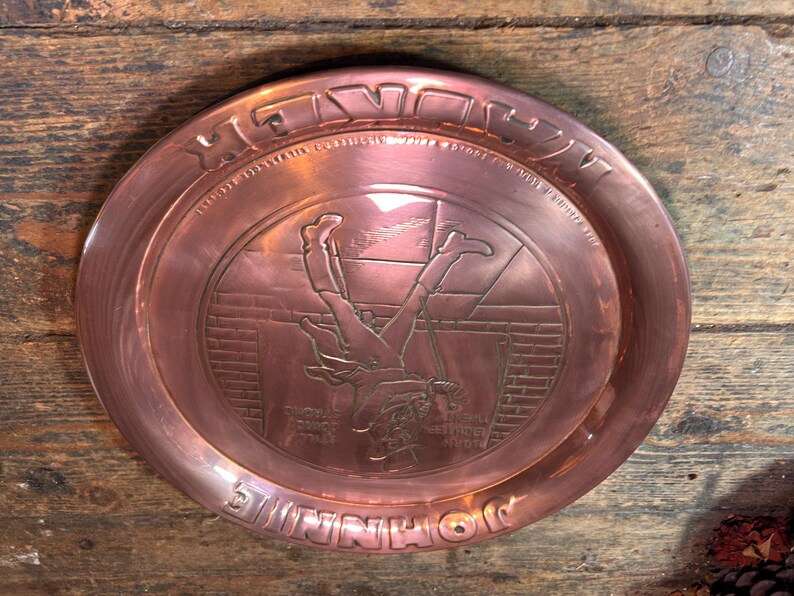 Puede incluir: Una bandeja de metal color cobre con un dise&ntilde;o en relieve. La bandeja presenta una imagen central de una figura cayendo, rodeada de texto. Las palabras "SHINNIHO" y "HE'S DOWN" son visibles. La bandeja es circular y tiene un borde elevado.