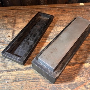 Puede incluir: Una piedra de afilar rectangular con una superficie metálica, colocada en una base de madera oscura. La piedra está junto a una bandeja de madera a juego. Los artículos están sobre una superficie de madera envejecida.