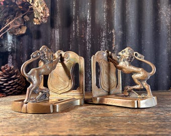 Puede incluir: Un par de sujetalibros antiguos de bronce. Cada sujetalibros presenta una figura de león con un respaldo en forma de escudo. Los leones están en una pose encabritada, con las patas apoyadas en el escudo. Los sujetalibros tienen una base rectangular.