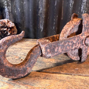 Peut inclure: Une poulie en métal rouillé antique avec un grand crochet. La poulie a une roue et un support, tous montrant des signes d'âge et d'usure. Le crochet est incurvé et robuste, suggérant qu'il était utilisé pour soulever des charges lourdes. L'article est posé sur une surface en bois.