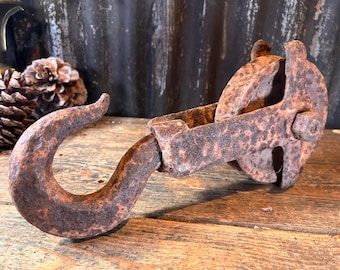 May include: An antique, rusty metal pulley with a large hook. The pulley has a wheel and a bracket, all showing signs of age and wear. The hook is curved and sturdy, suggesting it was used for heavy lifting. The item is set on a wooden surface.