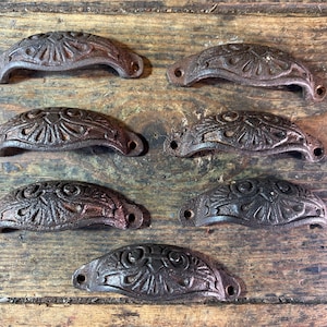 May include: Six antique-style cast iron drawer pulls with a dark brown finish. Each pull has a curved, half-moon shape with a detailed floral design. The pulls are arranged on a weathered wooden surface.