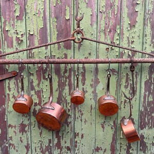 Puede incluir: Un antiguo colgador de ollas de cobre con seis ollas de cobre colgadas de ganchos. El colgador es de metal oxidado y está montado sobre una superficie de madera verde y marrón desgastada. Las ollas varían en tamaño.